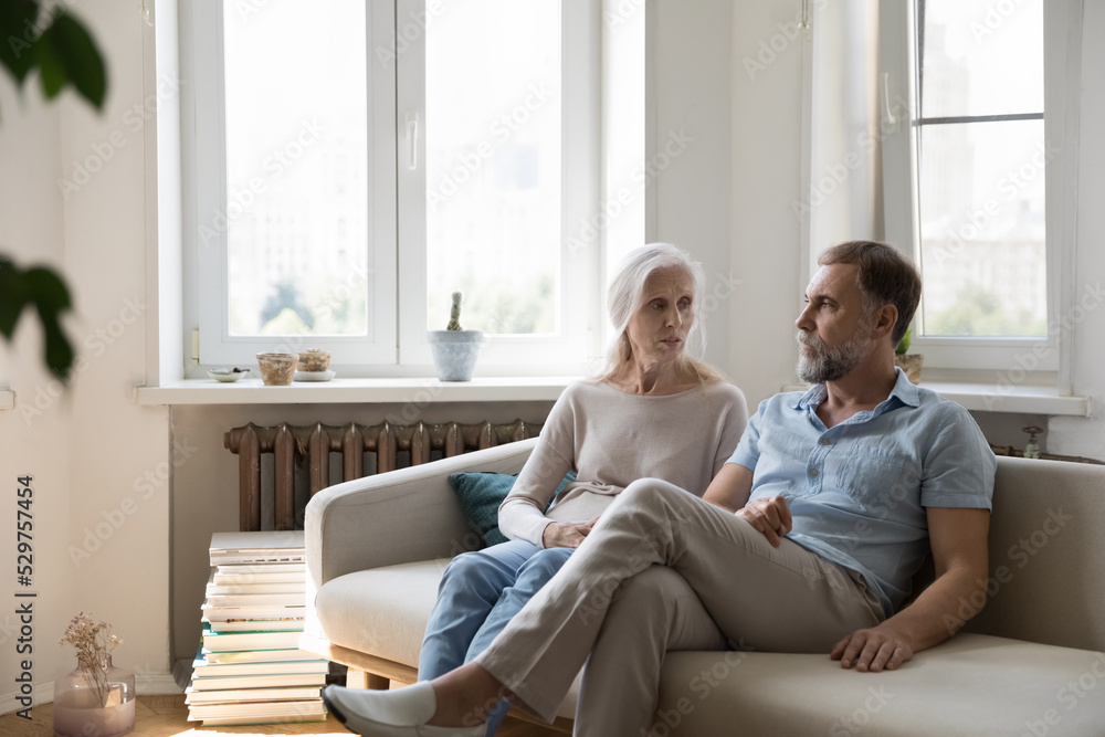Serious concerned senior couple talking on home couch, having relationship problems, discussing bad news. Mature older wife asking offended husband for forgiveness. Marriage crisis