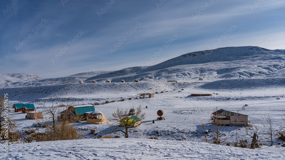 In the snow-covered valley, you can see the wooden houses of the campsite and spherical glamping tents. A mountain range against a background of blue sky and cirrus clouds. Altai. Kyzyl Chin In the snow-covered valley, you can see the wooden houses of the campsite and spherical glamping tents. A mountain range against a background of blue sky and cirrus clouds. Altai. Kyzyl Chin