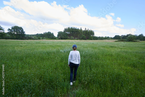 Rear view of woman wearing casual clothes and cap standing in front of green meadow under cloudy blue sky enjoying outdoors in nature.