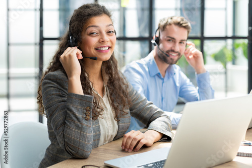 Female customer support operator with headset and smiling, with collegues at background.