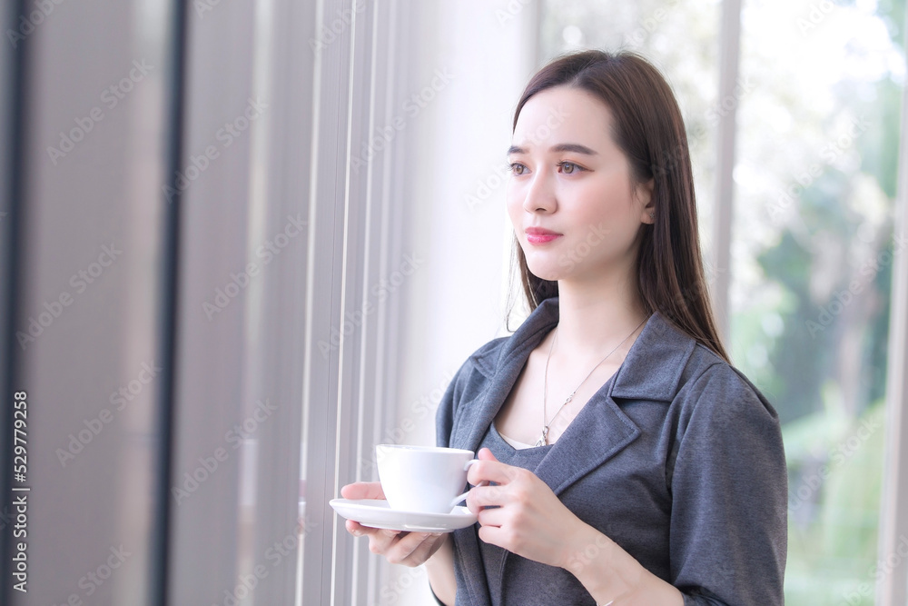Asian working woman is looking out of window to think something while holds coffee cup in her hand. Asian working woman is looking out of window to think something while holds coffee cup in her hand.