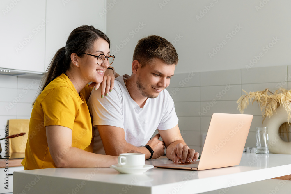 Pleasant married couple is sitting in a modern kitchen looking at a laptop screen. A young family is surfing the web, shopping online or booking flights. Pleasant married couple is sitting in a modern kitchen looking at a laptop screen. A young family is surfing the web, shopping online or booking flights.