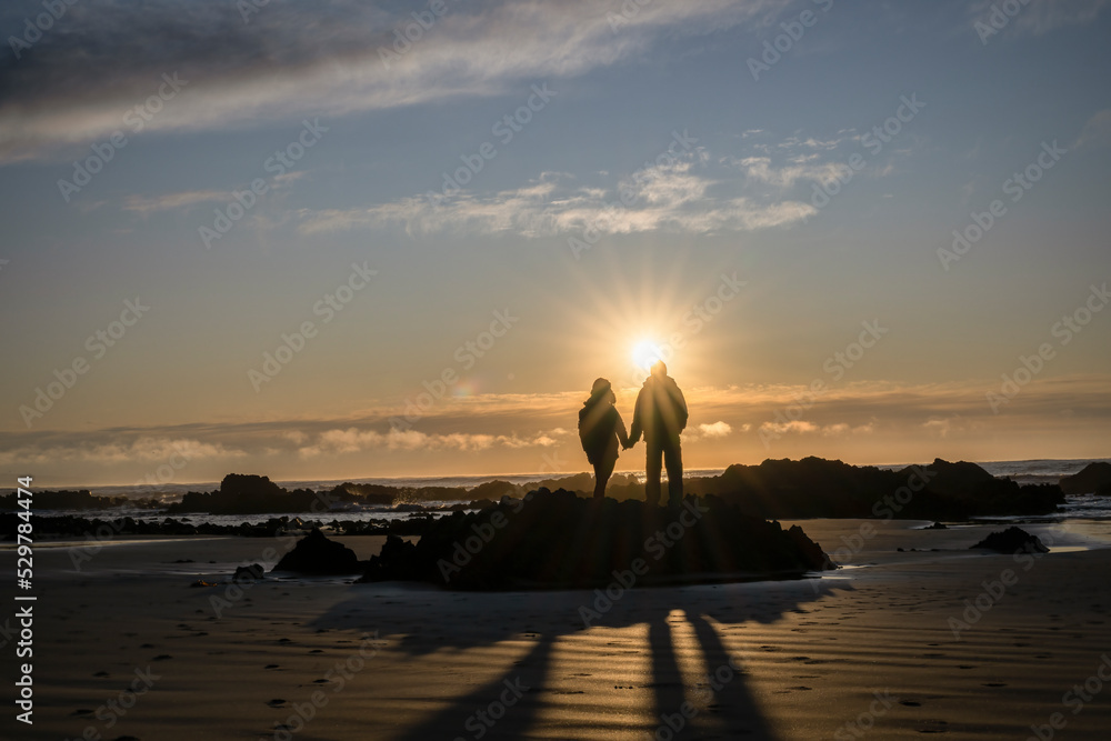Couple holding hands, watching the sunrise at a rocky beach.  Catlins, South Island.