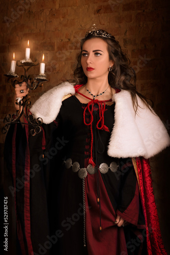 Fantastic medieval woman with a candlestick in her hands. Royal velvet vintage red dress with fur and gold, princess, queen, brunette with long hair. Castle, dusk, night, luxury