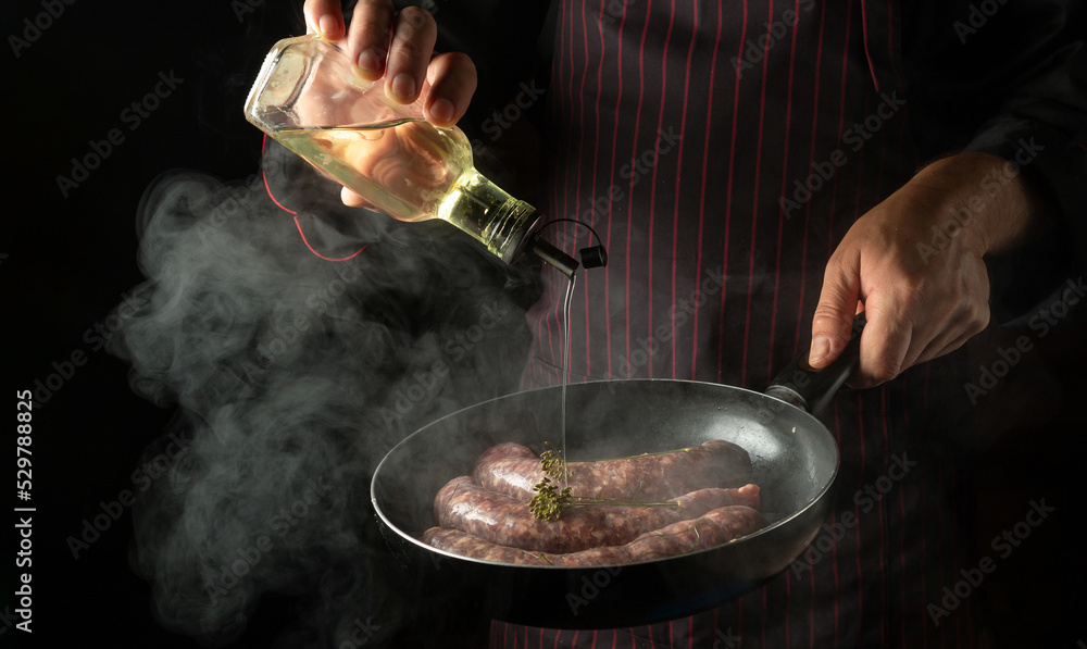 The chef adds oil to a hot frying pan with sausages. Menu or recipe concept for hotel on black background The chef adds oil to a hot frying pan with sausages. Menu or recipe concept for hotel on black background