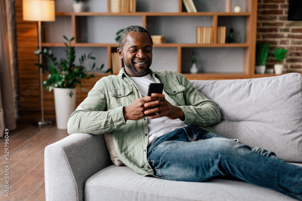 Glad mature african american male in casual resting and relaxing, chatting on smartphone, watching video