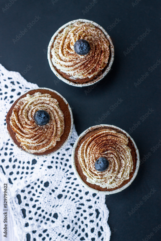 Three delicately delicious muffins with butter cream and blueberries on a lacy napkin on a dark table. Delicious dessert. Top view Three delicately delicious muffins with butter cream and blueberries on a lacy napkin on a dark table. Delicious dessert. Top view
