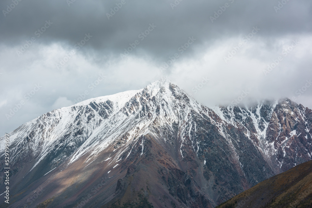 Atmospheric aerial view to sunlit high snow-white mountain with peaked top in rainy low clouds at changeable weather. Dramatic scenery with large snow mountain with pointed peak in low gray cloudy sky