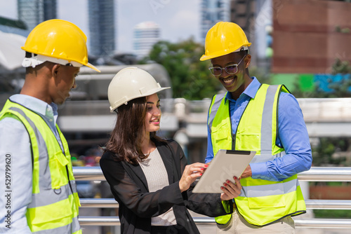 caucasian businesswoman in black suit wear helmet working on tablet at construction site outside building in city and standing near black engineer colleagues