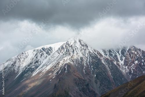 Atmospheric aerial view to sunlit high snow-white mountain with peaked top in rainy low clouds at changeable weather. Dramatic scenery with large snow mountain with pointed peak in low gray cloudy sky