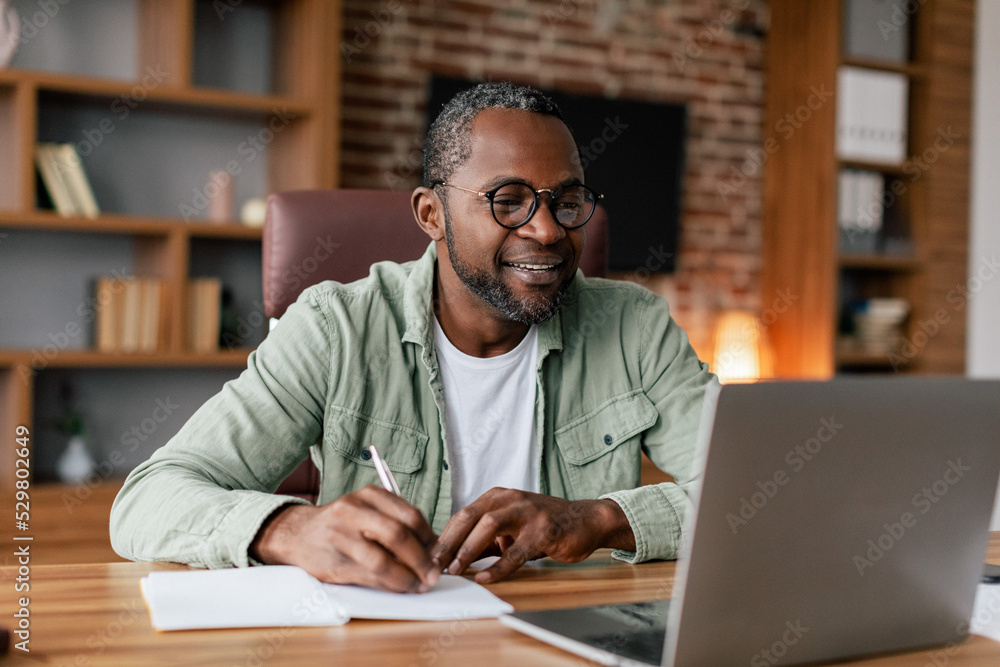 Smiling middle aged african american male in glasses and casual working on laptop in home office interior