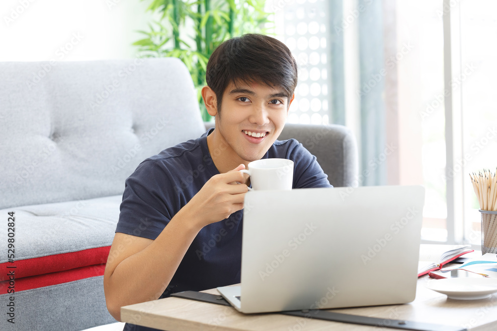 Happy Asian casual man working with laptop holding a cup of coffees, sitting on the floor in the house. Happy Asian casual man working with laptop holding a cup of coffees, sitting on the floor in the house.