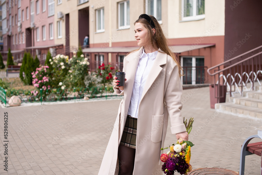 Fototapeta premium A cute girl in a coat holds a glass of coffee and a bouquet of flowers on the street