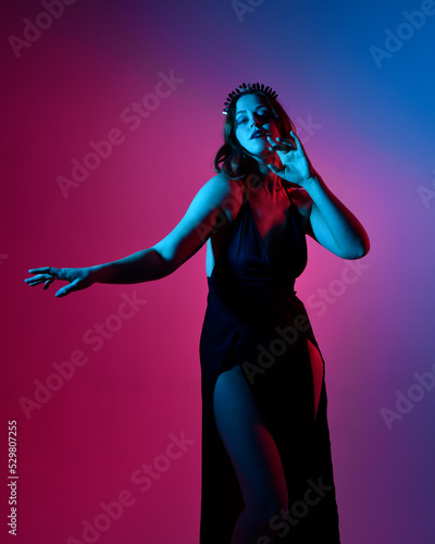 Close up portrait of beautiful woman model wearing elegant black dress and crown, posing against a studio background with fantasy inspired arm gestures, multi coloured creative lighting.