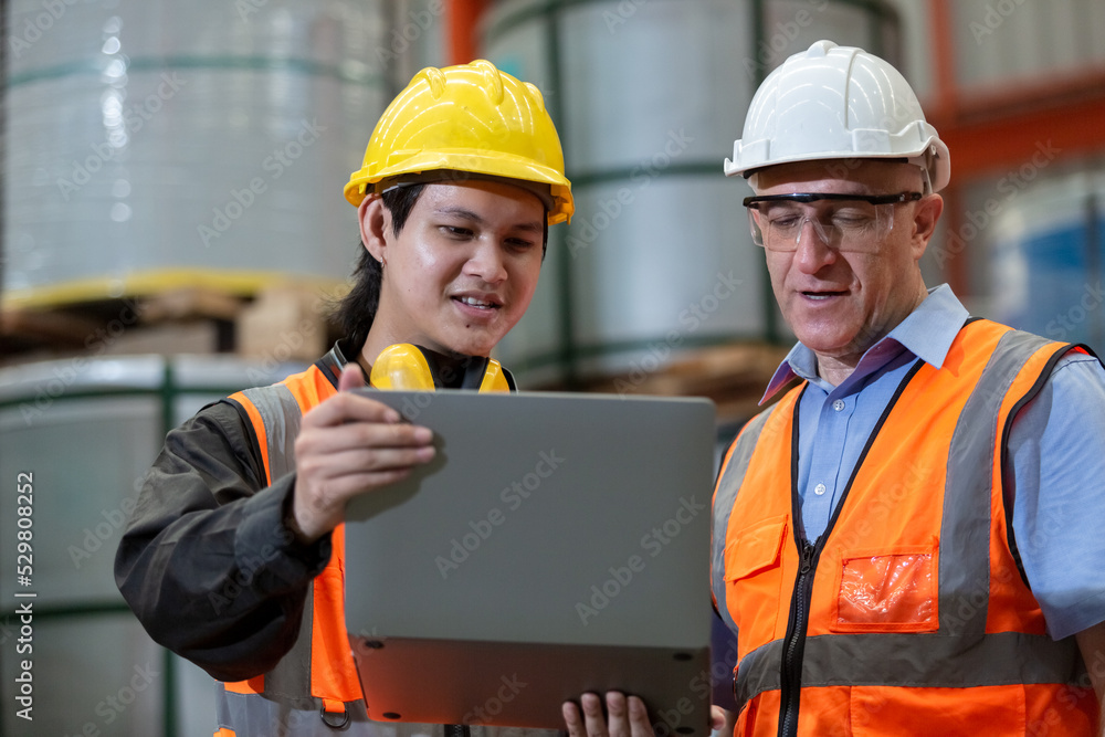 Two man industrial engineers wear hard hats and uniform using laptop learning project of factory inside heavy industry manufacturing. Supervisor teaching work to employee. Two man industrial engineers wear hard hats and uniform using laptop learning project of factory inside heavy industry manufacturing. Supervisor teaching work to employee.