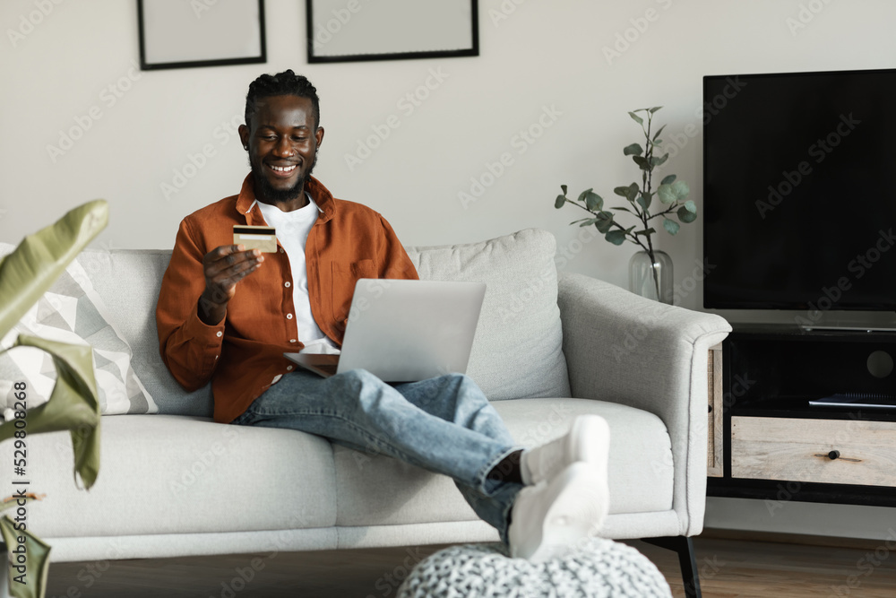 Excited african american man sitting on couch at home using laptop choosing what to buy and holding credit card