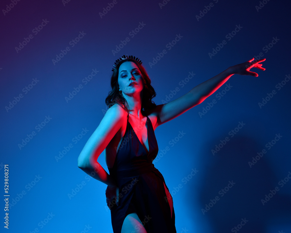 Close up portrait of beautiful woman model wearing elegant black dress and crown, posing against a studio background with fantasy inspired arm gestures, multi coloured creative lighting.

