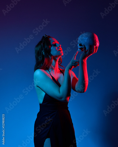 Close up portrait of beautiful woman model wearing elegant black dress and crown, posing against a studio background with fantasy inspired arm gestures, holding skull.multi coloured creative lighting. © faestock