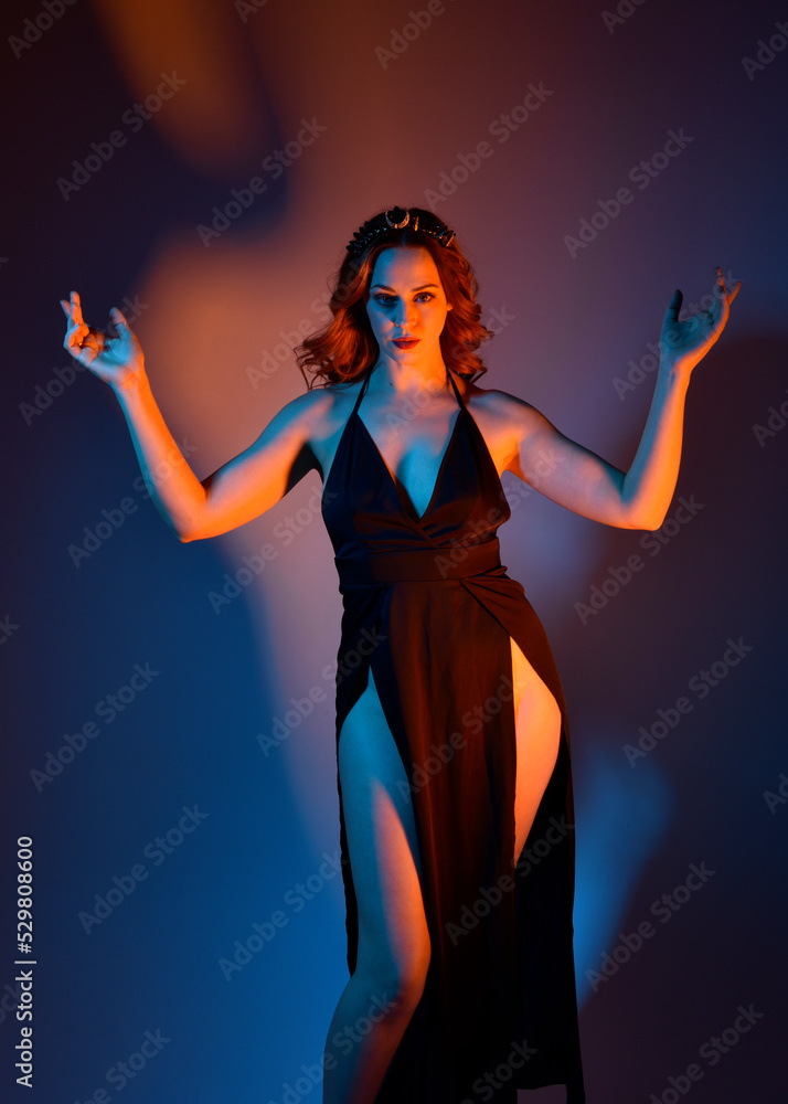 Close up portrait of beautiful woman model wearing elegant black dress and crown, posing against a studio background with fantasy inspired arm gestures, multi coloured creative lighting.
