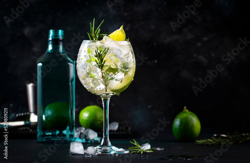 Gin tonic lime alcoholic cocktail drink with dry gin, rosemary, tonic and ice in big wine glass. Black bar counter background, steel bar tools photo