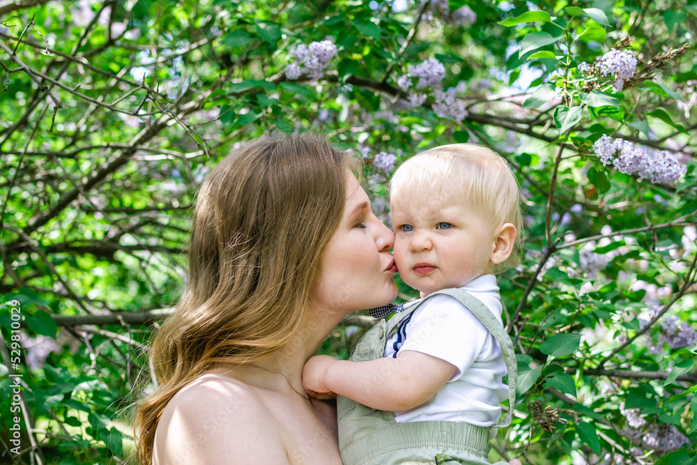 Young beautiful mother is kissing her little son in sunlight on background of blooming lilac trees.