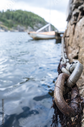Iron chain on old stone pier, sea waves and moored sailboat and rocky shore out of focus. Bottom view.
