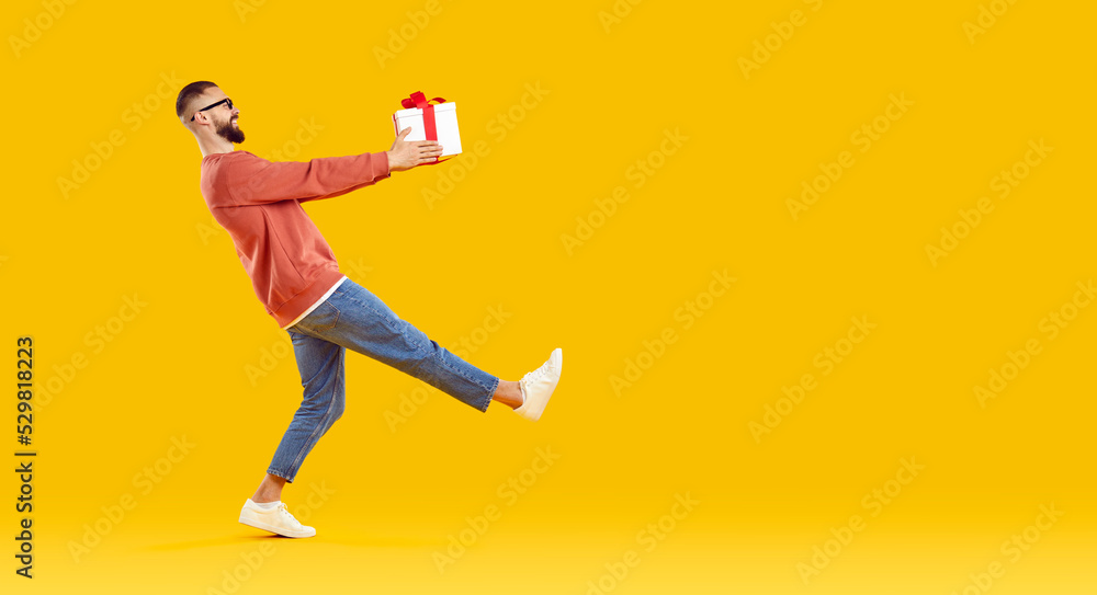 Obraz premium Happy man carrying a gift box. Side view of a funny young guy in casual clothes holding a present and walking against a bright orange yellow blank empty copy space studio background. Holiday concept