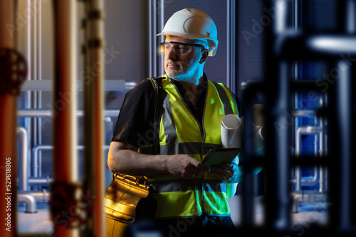 Boiler room engineer. Man among pipes. Human in engineers clothes is standing indoor. Guy in room with utilities. Boiler room design concept. Worker with tablet among boiler equipment. Art focus.