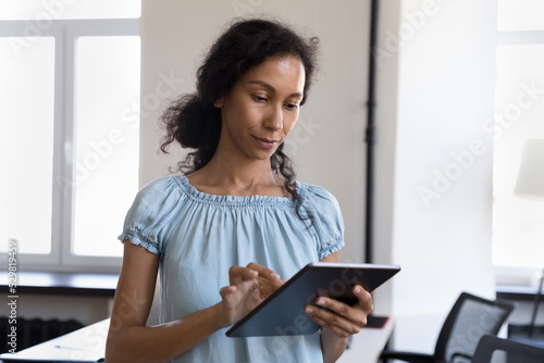 Serious young African employee woman in casual using professional online app on tablet, reading text on gadget screen, browsing internet, watching video content. Business communication concept