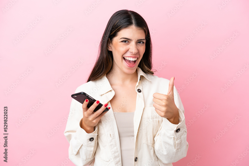 Young Italian woman isolated on pink background using mobile phone while doing thumbs up Young Italian woman isolated on pink background using mobile phone while doing thumbs up
