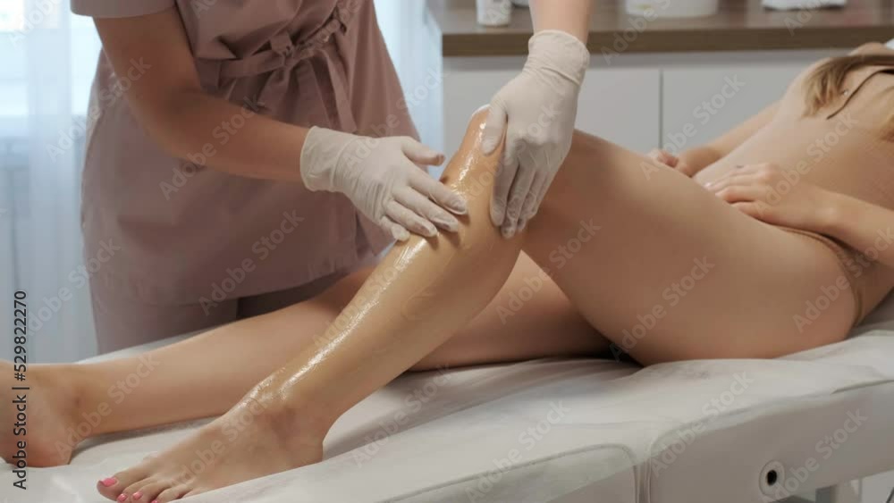 A female depilatory expert applies sugar paste on the patient's leg. The process of hair depilation on the girl's leg with sugar paste by a professional master.