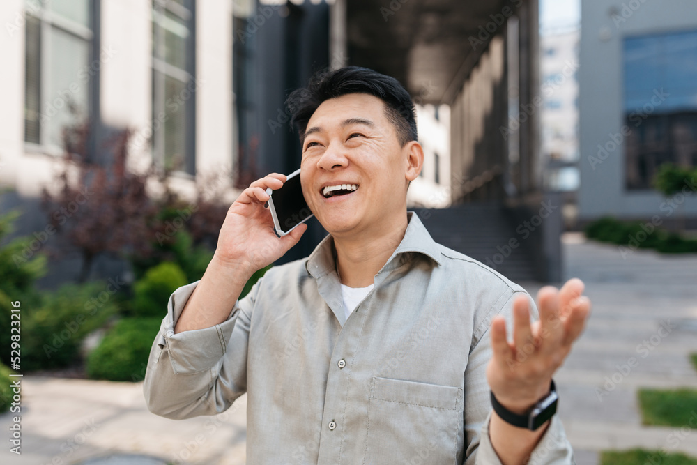 Cheerful asian middle aged businessman calling by phone outdoors, walking near modern office building Cheerful asian middle aged businessman calling by phone outdoors, walking near modern office building