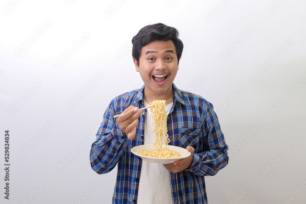 Portrait of happy Asian man in blue plaid shirt eating delicious instant noodles with chopsticks served on plate. Isolated image on white background Portrait of happy Asian man in blue plaid shirt eating delicious instant noodles with chopsticks served on plate. Isolated image on white background