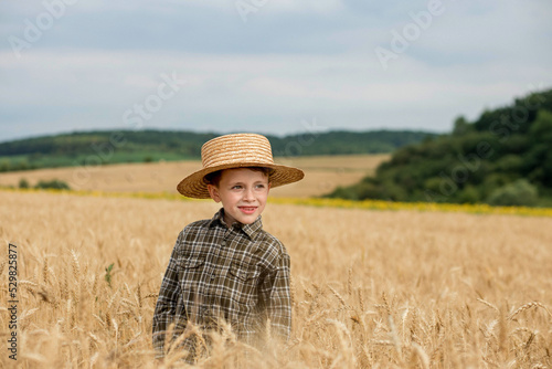 A smiling little farmer boy in a plaid shirt and straw hat poses for a photo in a wheat field. Heir of farmers © volody10