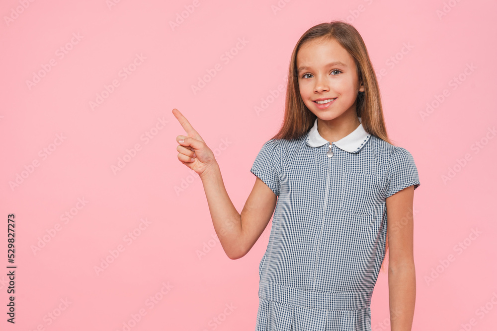 Happy preteen teenage schoolgirl in blue dress looking at camera showing pointing at copyspace isolated in pink background Happy preteen teenage schoolgirl in blue dress looking at camera showing pointing at copyspace isolated in pink background