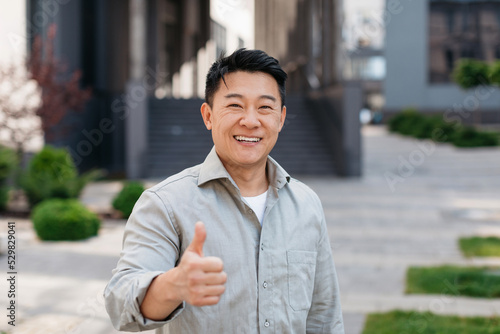 Portrait of positive asian man standing outdoors and gesturing thumb up, walking in urban city area, copy space photo