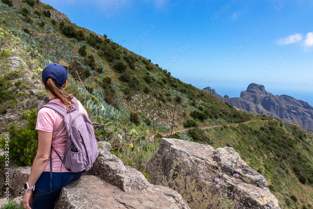 Rear view on woman sitting on rock in Teno mountain massif, Tenerife, Canary Islands, Spain, Europe. Coastal hiking trail leading to remote village Masca. View on rock formations and Atlantic Ocean