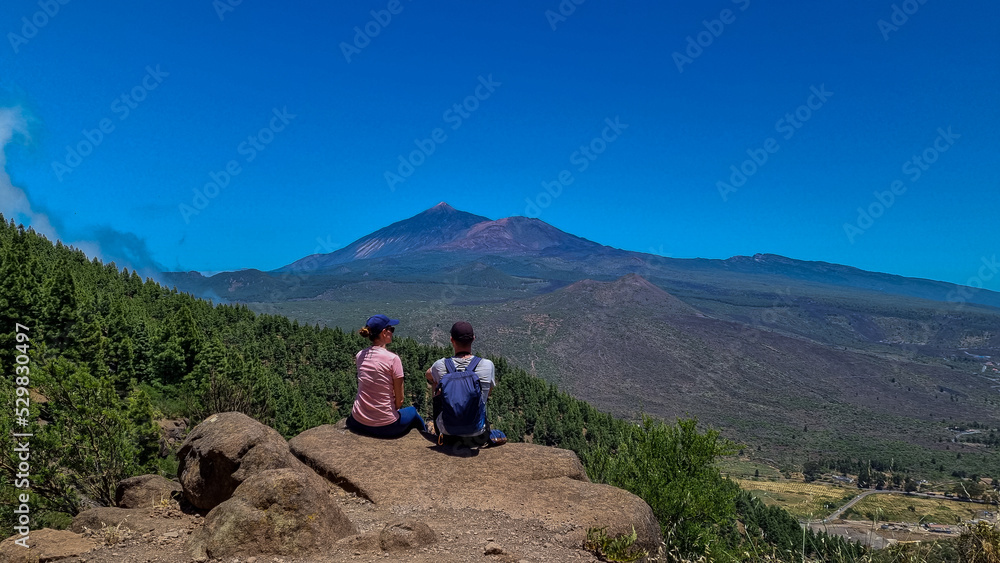 Sitting couple with scenic view on volcano Pico del Teide surrounded by pine tree forest, Teno mountain, Tenerife, Canary Islands, Spain, Europe. Hiking trail from Santiago to Masca via Pico Verde