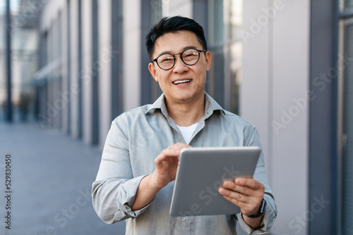Happy asian middle aged businessman using tablet, looking and smiling at camera, standing near modern office building