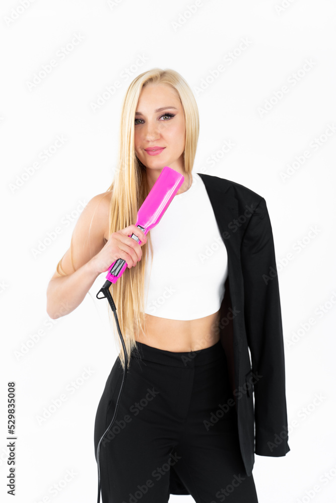 In a white room, a stylish blonde woman with long hair in black straightens her long blonde hair with a pink curling iron In a white room, a stylish blonde woman with long hair in black straightens her long blonde hair with a pink curling iron