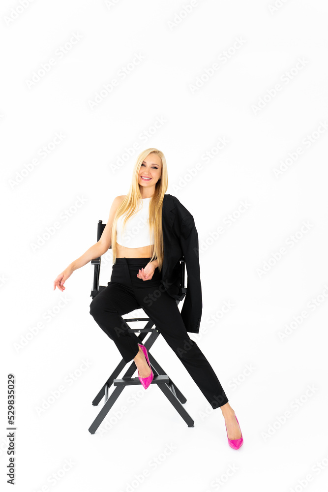 One young European girl in black clothes and pink shoes with long tousled blond hair with a smile on her face is sitting on a black director's chair in a white room One young European girl in black clothes and pink shoes with long tousled blond hair with a smile on her face is sitting on a black director's chair in a white room