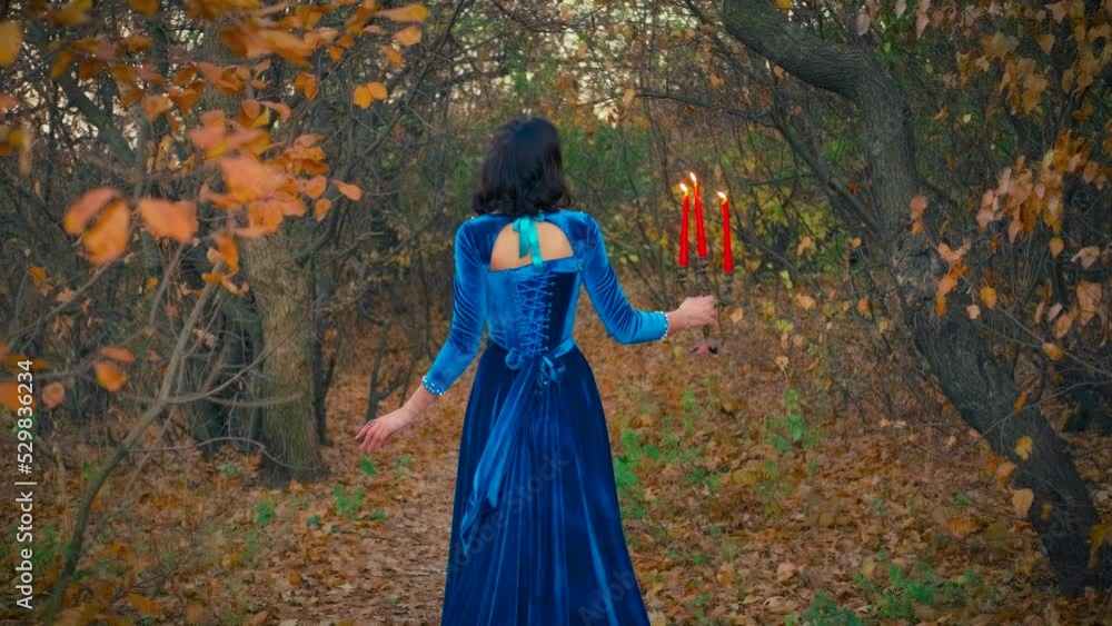 Beauty autumn forest bare black tree trunks. Gothic elegant fantasy woman queen. girl vampire princess turned away, holding candlestick red candles burning fire. Long blue vintage dress back rear view Beauty autumn forest bare black tree trunks. Gothic elegant fantasy woman queen. girl vampire princess turned away, holding candlestick red candles burning fire. Long blue vintage dress back rear view