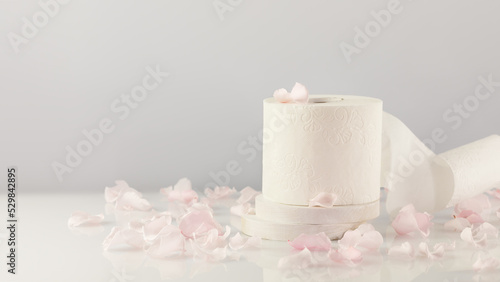 Roll of white toilet paper on a white table with rose petals and copy space. White toilet tissue, Hygiene product. Restroom soft touch toilet paper. Soft focus style image, horizontal orientation