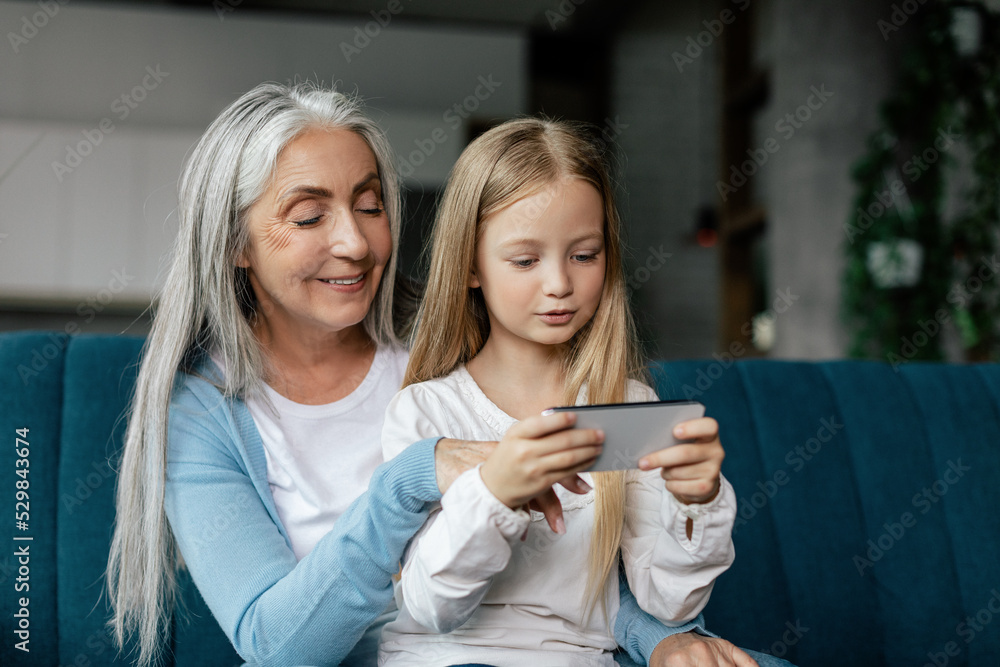 Busy glad caucasian little girl and elderly granny watch video, enjoy online game on smartphone, use new app Busy glad caucasian little girl and elderly granny watch video, enjoy online game on smartphone, use new app