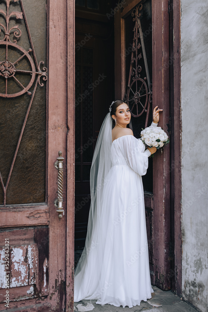A beautiful bride in a white long dress stands near an old wooden door outdoors with a bouquet of roses in her hands. Wedding portrait, photography.