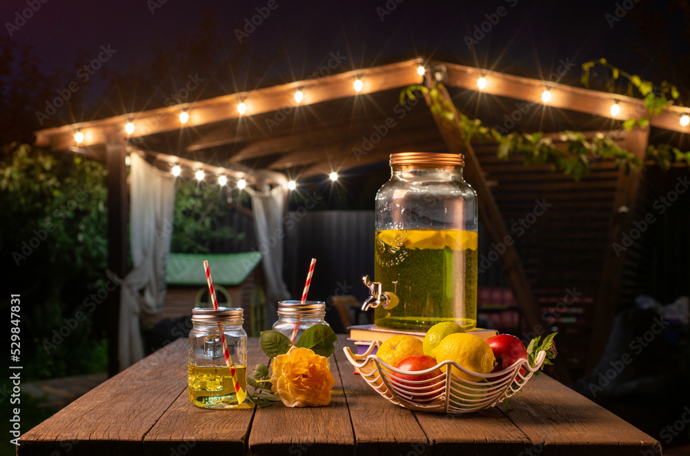 drink station with small bottles and homemade lemonade on wooden terrace with abstract night light bokeh of night festival in garden, copy space for display of product or object presentation