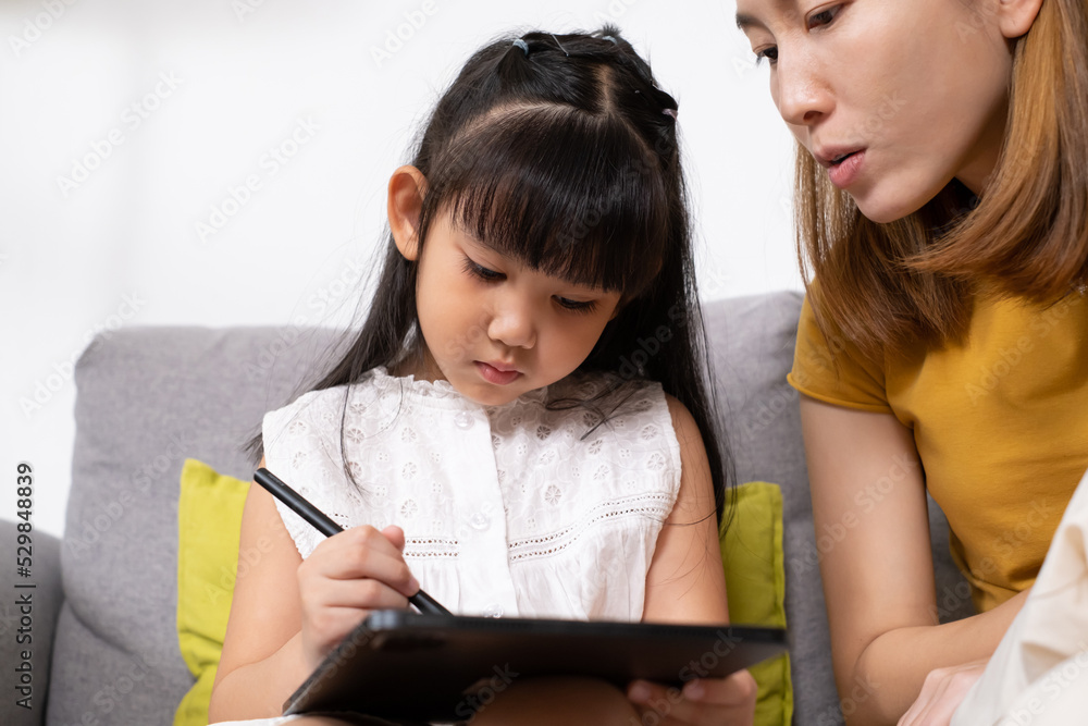Young Asian girl using digital tablet for painting picture or study online lesson at home, with mother support and teaching the lesson inside of living room. Online education or home school concept. Young Asian girl using digital tablet for painting picture or study online lesson at home, with mother support and teaching the lesson inside of living room. Online education or home school concept.