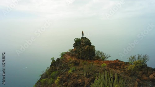 Fearless woman enjoys nature landscape on mountain top. Tourist stands on the edge of a cliff at great height. Blue ocean water below. Peak of rock. Drone flight over the summit. photo