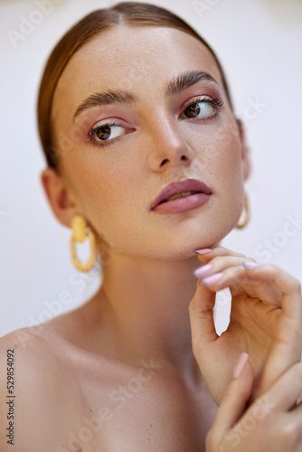 Female model with red hair, brown eyes, freckled skin, poses for a beauty shoot. She wears gold earrings and has her hand to her face.