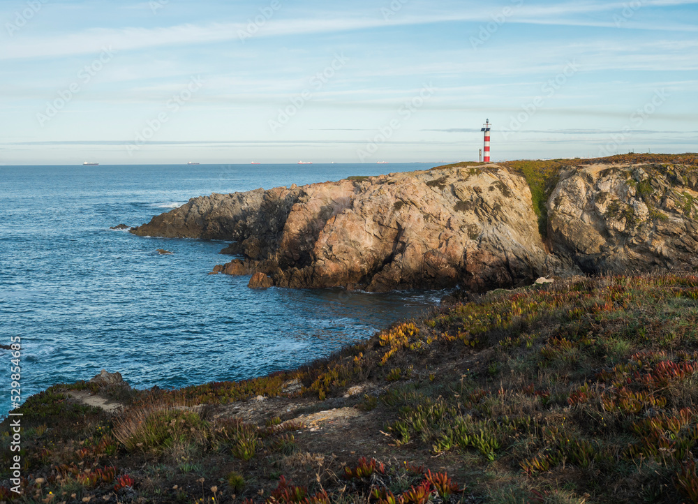 View of the sea and cliffs with the red and white lighthouse at praia pequena beach at wild Vicentina coast in Porto Covo, Portugal. Golden hour.
